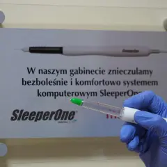 Ręka w niebieskiej rękawiczce trzyma aplikator z bupiwakainą, obok tablica informująca o bezbolesnym znieczuleniu komputerowym SleeperOne.