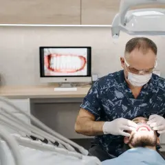 Dentysta w masce i rękawiczkach wykonuje zabieg, który może pomóc w usuwaniu kamienia nazębnego w domu. Na ekranie widać zbliżenie zębów.