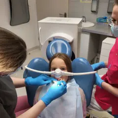 Dziecko u dentysty z maską do podawania gazu rozweselającego. Dwie higienistki dbają o komfort małego pacjenta podczas zabiegu.