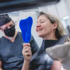 Uśmiechnięta pacjentka ogląda swoje zęby w lusterku, podczas gdy dentysta w masce obserwuje. Czy po wyrwaniu zęba można lecieć samolotem? Zapytaj specjalistę.
