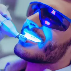 Mężczyzna w okularach ochronnych przechodzi zabieg wybielania zębów u dentysty. Dowiedz się, jaka jest wybielanie zębów u dentysty cena.