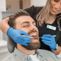 Uśmiechnięta dentystka bada zęby pacjenta, który lekko zaciska skurcz szczęki, by ułatwić badanie.