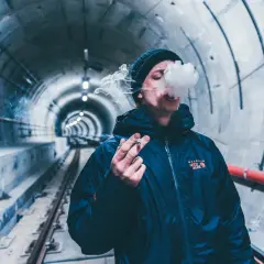 Młody mężczyzna w czapce i kurtce pali papierosa w tunelu metra. Dym unosi się wokół jego twarzy, jakby to był jeden papieros po wyrwaniu zęba.