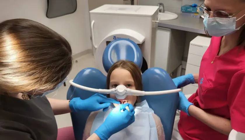 Dziecko u dentysty z maską do podawania gazu rozweselającego. Dwie higienistki dbają o komfort małego pacjenta podczas zabiegu.