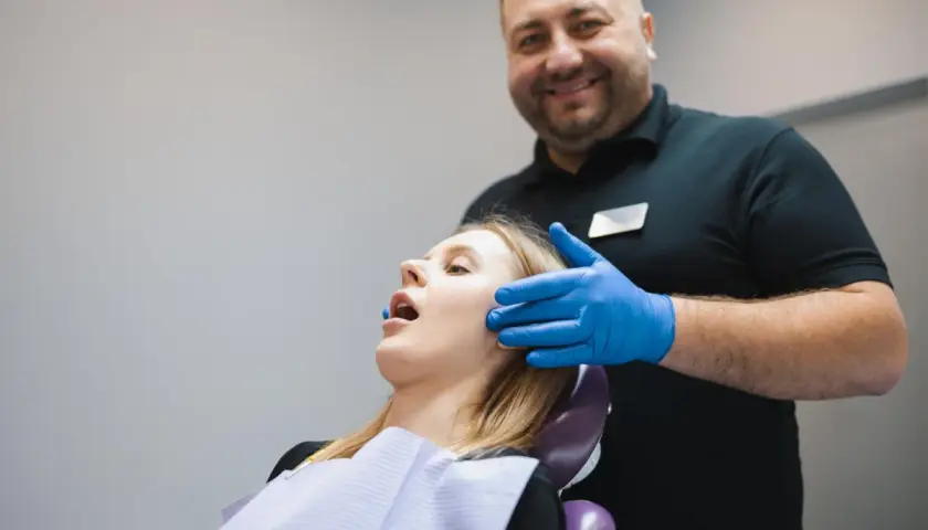 Uśmiechnięty dentysta w niebieskich rękawiczkach bada pacjentkę. Zespół Costena dba o zdrowie jamy ustnej.