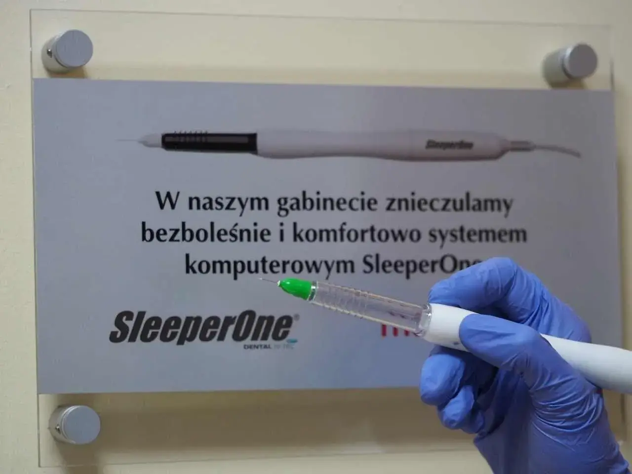 Ręka w niebieskiej rękawiczce trzyma aplikator z bupiwakainą, obok tablica informująca o bezbolesnym znieczuleniu komputerowym SleeperOne.