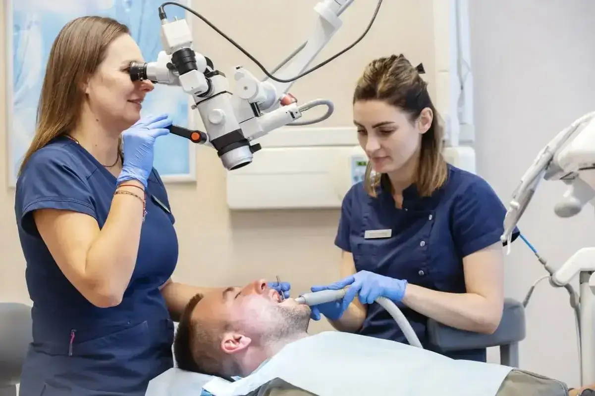 Dentystka znieczula pacjenta przewodowo, używając mikroskopu do precyzyjnej pracy. Asystentka przygotowuje narzędzia.